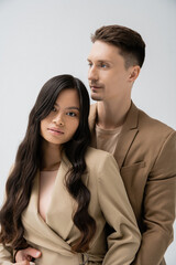 trendy man embracing pretty asian woman with long brunette hair isolated on grey.