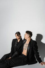 tattooed man in black fashionable suit looking at elegant asian woman sitting on grey background.