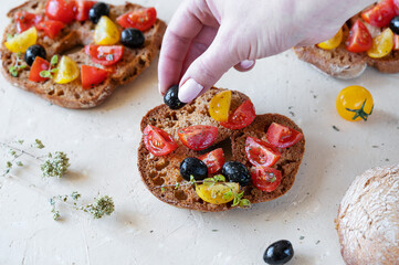 Friselle are typical Italian bread topped with red and yellow tomatoes, olive oil, olives and oregano on light sand background. Woman hand is garnishing Friselle with olive