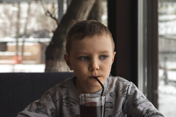 Adorable little kid boy is sitting at a table in a cafe and drinking a juice from a glass through a straw
