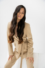 young asian woman with long brunette hair wearing beige suit and looking at camera near cube on white background.