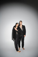 full length of barefoot multiethnic couple in black stylish pantsuits standing on grey background.