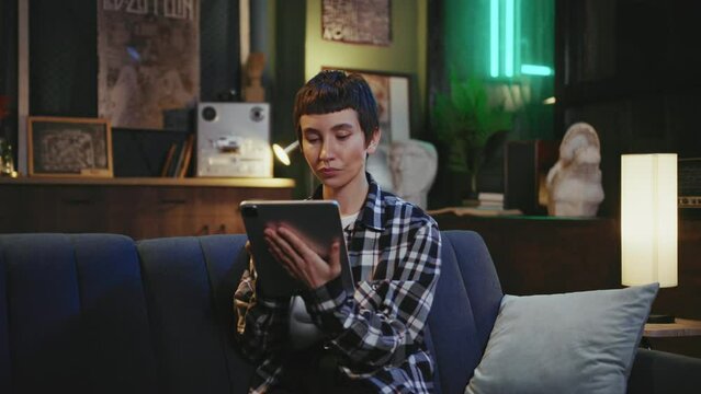 Young serious focused woman with stylish hairstyle sitting on sofa in cozy loft office. Attractive girl concentrated on work looking at screen of digital tablet. Modern interior with neon light
