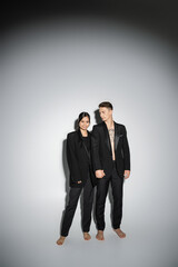 full length of barefoot interracial couple in black elegant pantsuits holding hands on grey background.