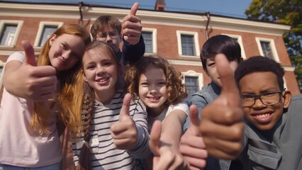 Portrait of group of diverse school kids showing thumbs up outside school. Realtime