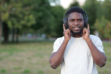African American man wearing black headphones listens to favourite music. Bearded guy dances to excited music standing in green park, copysapce