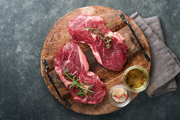 Raw beef steak. Marbled raw fresh Ribeye steak with rosemary, salt and pepper on cutting board on dark concrete background. Raw beef steak and spices for cooking. Mock up.
