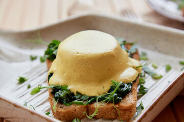 Vegan eggs benedict dish with wholegrain bread, creamy plant-based hollandaise sauce, smoked tofu, young onion, parsley, greens, pea sprouts.