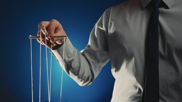 A businessman in a gray shirt and black tie controls a puppet with a wooden manipulator and strings. Hand handling at puppet by pulling strings to make the character move. Blue background. Close up.