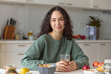 Smiling tanned Arabic Woman Enjoying Healthy Meal at Home. A happy woman with curly brown hair smiles looking at the camera in a domestic kitchen, surrounded by healthy food and household equipment. 