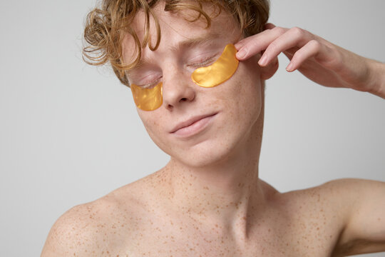 A portrait of a young man with red hair and perfect skin with freckles, shot in the studio against a grey background. The model poses and wears gold coloured gel eye patches. Young and beauty concept.