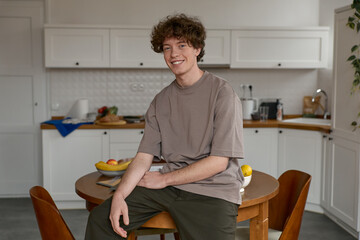 Portrait of a happy man with brown curly hair wearing casual clothing  looking at the camera and smiling while sitting on a table in modern white kitchen and enjoying life indoors. Lifestyle concept.