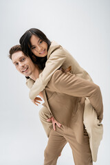 happy and stylish man piggybacking young asian woman while smiling at camera isolated on grey.