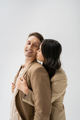 brunette asian woman embracing cheerful man wearing beige blazer isolated on grey.