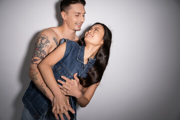 shirtless tattooed man hugging happy asian woman in denim vest on grey background.