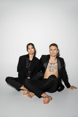 full length of barefoot interracial couple in black elegant pantsuits sitting with crossed legs on grey background.