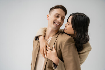 brunette asian woman embracing happy man in beige stylish blazer isolated on grey.