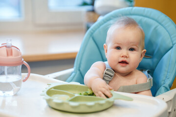 The kid is sitting in a high chair in the kitchen and trying to take food from the plate with his hands. Complementary food