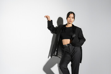 fashionable asian woman in black crop top and pantsuit looking at camera on grey background with...