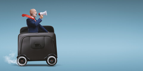 Businessman with megaphone riding a briefcase with wheels