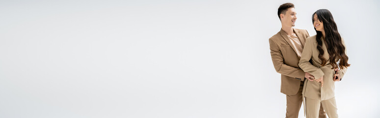 happy and stylish interracial couple in beige suits smiling at each other isolated on grey, banner.