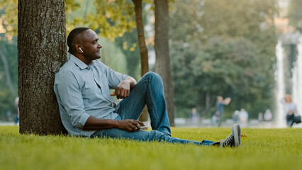 Happy calm relaxed elderly middle-aged male listening music with earphones mature senior adult African American ethnic man in wireless headphones sitting on grass in city park enjoy audio song sound