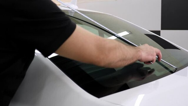 A male car mechanic measures the rear windshield with a measuring tape before gluing the tinting film.