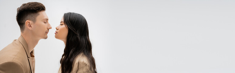 side view of long haired asian woman and trendy man posing face to face with closed eyes isolated on grey, banner.