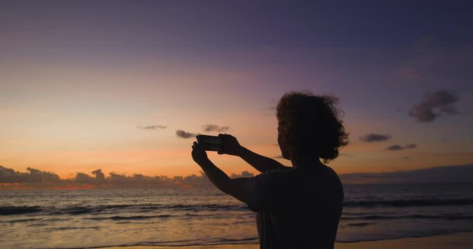 A female tourist on sea beach takes a selfie in the beautiful soft dawn light. Caucasian curly woman on vacation at sea takes selfies, photos of herself on beach early in the morning in the dawn sun