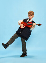 Happy kid, little musician wearing big size clothes posing with violin isolated over blue...