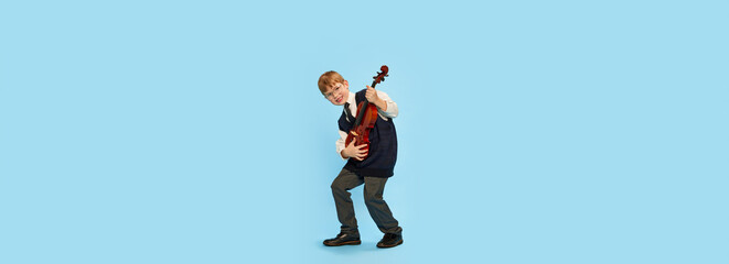 Happy kid, little musician wearing big size clothes posing with violin isolated over blue background. Education, music, classic art, new vision, fun concept © Lustre Art Group 