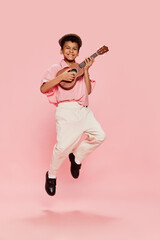 Happy emotional little african boy in retro style clothes and cap posing with ukulele guitar isolated on pink background.