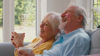 Retired senior couple with hot drinks sitting on sofa in lounge at home together watching off camera TV and laughing - shot in slow motion - Powered by Adobe