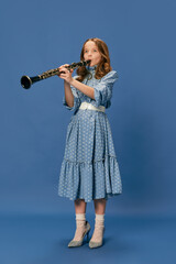 Monochrome portrait of charming nice little girl in dress and big shoes playing on clarinet over blue background. Music, fashion, hobby concept