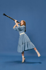 Monochrome portrait of charming nice little girl in dress and big shoes playing on clarinet over blue background. Music, fashion, hobby concept