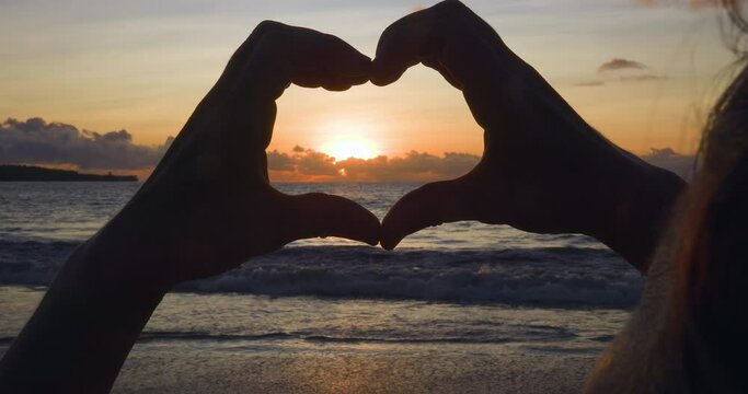 Silhouette of hands at sunset on seascape. Dark silhouette of women's hands that show heart against background of sea surf, waves, orange sun hiding behind horizon. Hand gesture enclosing sun in heart