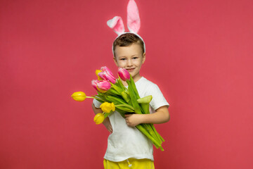 Little boy with easter bunny ears holds a bouquet of tulips on pink studio background. Copy space