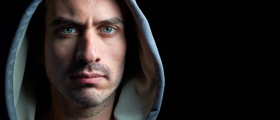 Panoramic horizontal close up portrait of young handsome hooded man with blue eyes looking at the camera with serious tranquil expression isolated on dark black background with copy space. - Powered by Adobe