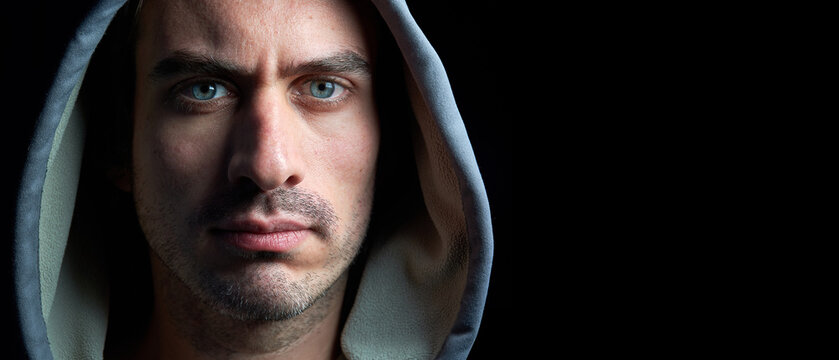 Panoramic Horizontal Close Up Portrait Of Young Handsome Hooded Man With Blue Eyes Looking At The Camera With Serious Tranquil Expression Isolated On Dark Black Background With Copy Space.