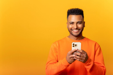 Photo of handsome cheerful smiling man holding mobile phone in the hand and smiling, isolated over yellow background, chatting using smartphone in front camera