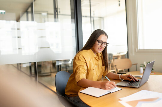 Smiling female student using laptop for watching webinars, lectures, sits at desk in office and taking notes, woman taking a part in online educational course, researching topic