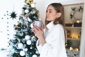 Young woman with cup of hot drink near Christmas tree