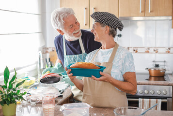 Cheerful senior woman holding her freshly baked homemade plumcake at her husband washing the...