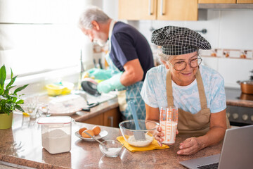 Happy elderly couple working together in the home kitchen, husband washing dishes, wife preparing a homemade cake looking at web pages by laptop for the right recipe