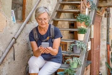 Attractive senior tourist woman sitting on a wooden staircase visiting an old swiss village typing...