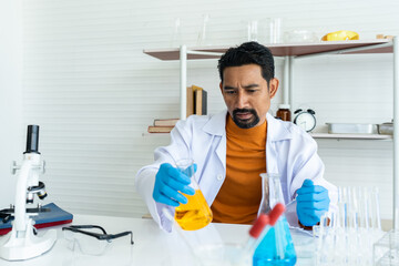 A male teacher in white lab coat with rubber gloves with many laboratory tools on shelves and table. Picking yellow chemical flask on table for checking carefully. Before chemistry classroom begin..