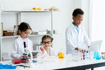 Two little Asian girls and one young African boy in white lab coat with many laboratory tools on shelves and table. Help each other for experiment class by checking and recording result information.