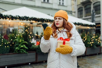 Worried concerned girl having problems, looking at smartphone screen with puzzled face on winter street. Winter holidays sale. Scam, fraud concept. Upset young woman using phone and reading bad news