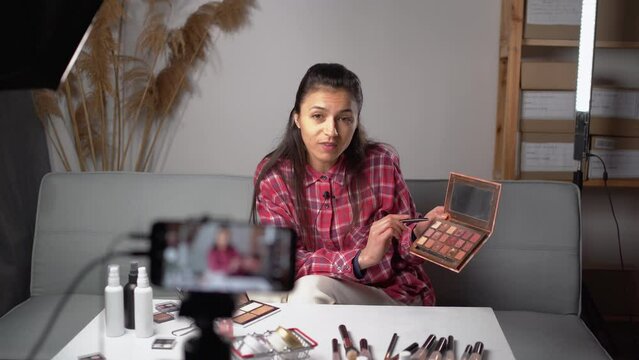 Beauty blogger or vlogger woman filming daily make-up routine tutorial and showing eye shadow palette near camera on tripod. Influencer girl live streaming cosmetics product review in home studio.