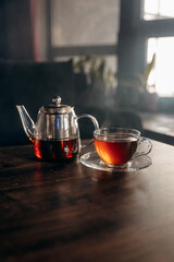 Close-up photo of a transparent teapot filled with tea and a transparent glass cup standing on a table with smoke. Hot tea for health. Smoky atmosphere for tea